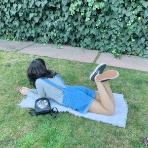 May include: A person wearing denim overalls and a grey long-sleeved shirt is lying on a pink and white plaid blanket. They are wearing black and white sneakers and have a black heart-shaped backpack with colorful accents.