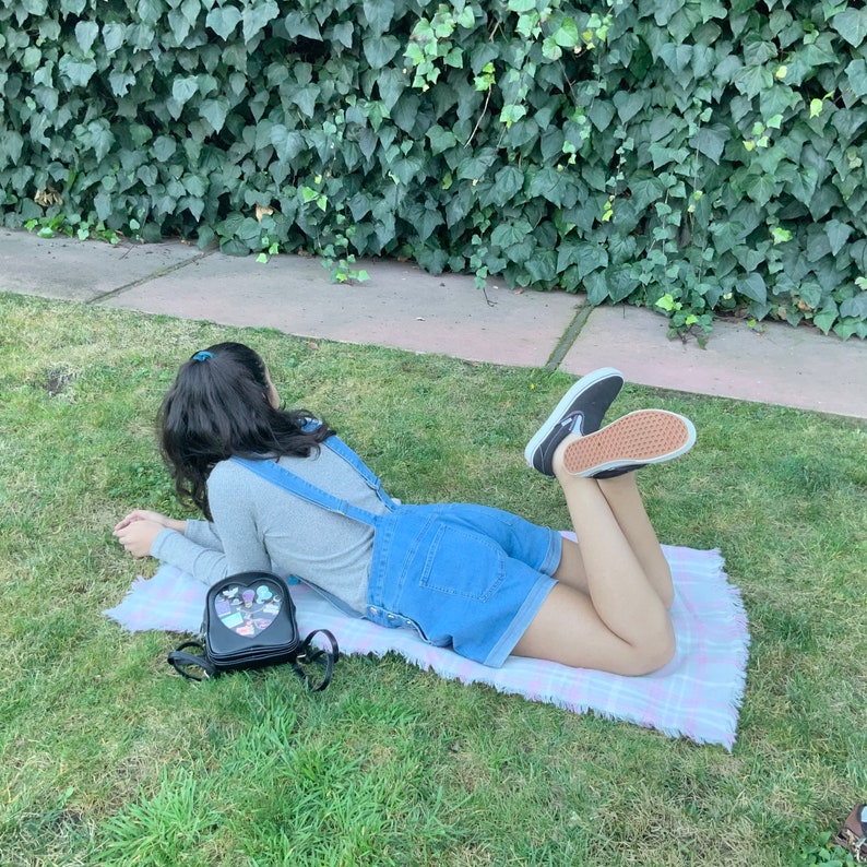 May include: A person wearing denim overalls and a grey long-sleeved shirt is lying on a pink and white plaid blanket. They are wearing black and white sneakers and have a black heart-shaped backpack with colorful accents.