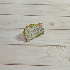 May include: A gold-colored enamel pin with a white background and green and pink floral accents. The pin says "Happy Introvert" in a cursive font.