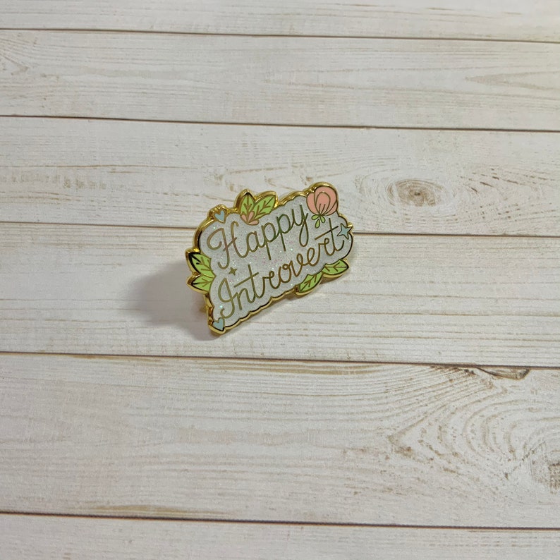 May include: A gold-colored enamel pin with a white background and green and pink floral accents. The pin says "Happy Introvert" in a cursive font.