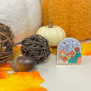 Op de afbeelding: Een gouden emaille pin met een vensterscène met een vrouw die bij een raam zit en uitkijkt op een kleurrijk herfstlandschap. De pin heeft een witte achtergrond en een gouden frame.