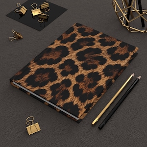 Può includere: Un taccuino con copertina rigida caratterizzato da un motivo leopardato marrone e nero, accompagnato da una matita color oro, una matita nera e diverse clip in metallo dorato disposte attorno al taccuino su una superficie scura e ruvida.
