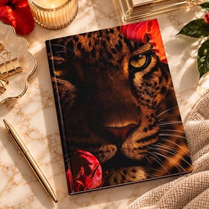 Può includere: Un diario rettangolare con copertina rigida caratterizzato da una stampa in primo piano ad alta risoluzione di un muso di leopardo con intensi occhi gialli, circondato da vivaci fiori di ibisco rossi. La copertina mostra dettagli del pelo.