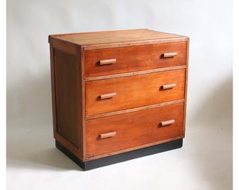 Puede incluir: Una cómoda de madera con tres cajones horizontales y tiradores de madera rectangulares sencillos. La pieza tiene un acabado de veta de madera marrón cálida y se apoya sobre una base negra sólida, proporcionando estabilidad a la unidad.