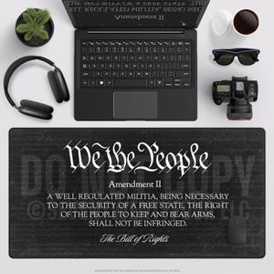 May include: A black desk setup featuring a laptop, headphones, sunglasses, a camera, and a mouse. A mousepad with the text "We the People" and the Second Amendment is the focal point. A small potted plant and a cup of coffee complete the scene.