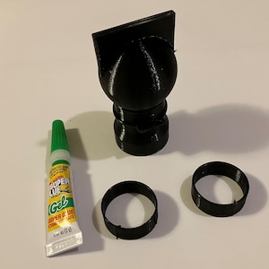 May include: A black 3D-printed object resembling a graduation cap, with two black rings and a tube of Super Glue Gel. The glue tube is white, yellow, and green, with text that reads "Super Glue Gel".
