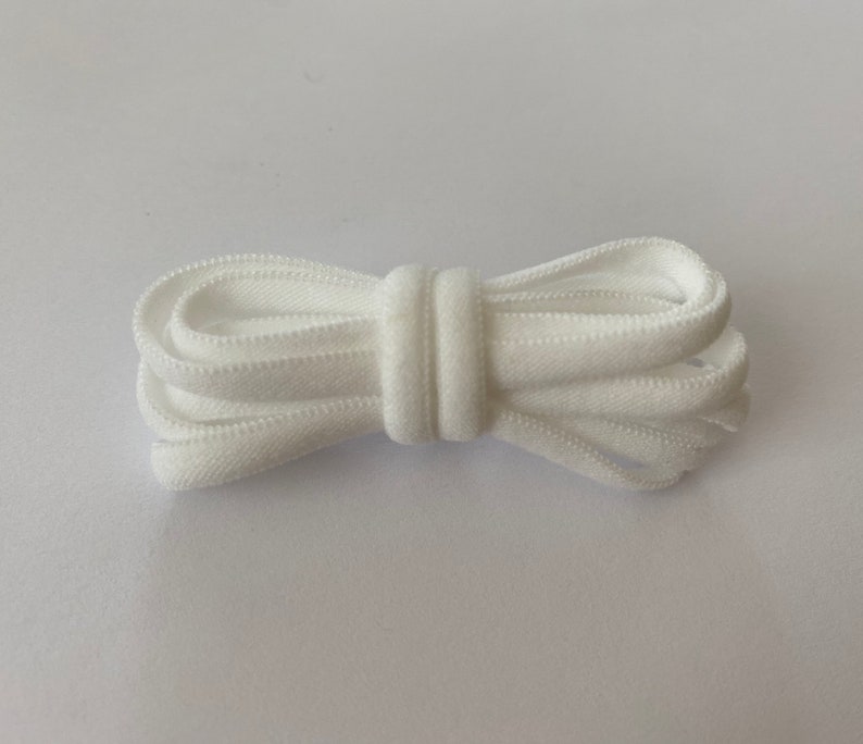 Pode incluir: Cord&atilde;o el&aacute;stico branco, dobrado ao meio, com textura lisa. O cord&atilde;o tem aproximadamente 1,2 cm de largura.