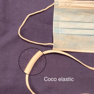 Pode incluir: Close-up de uma banda el&aacute;stica bege, tamb&eacute;m conhecida como "el&aacute;stico coco", sobre um fundo de tecido roxo. A banda el&aacute;stica &eacute; grossa e tem uma textura lisa.