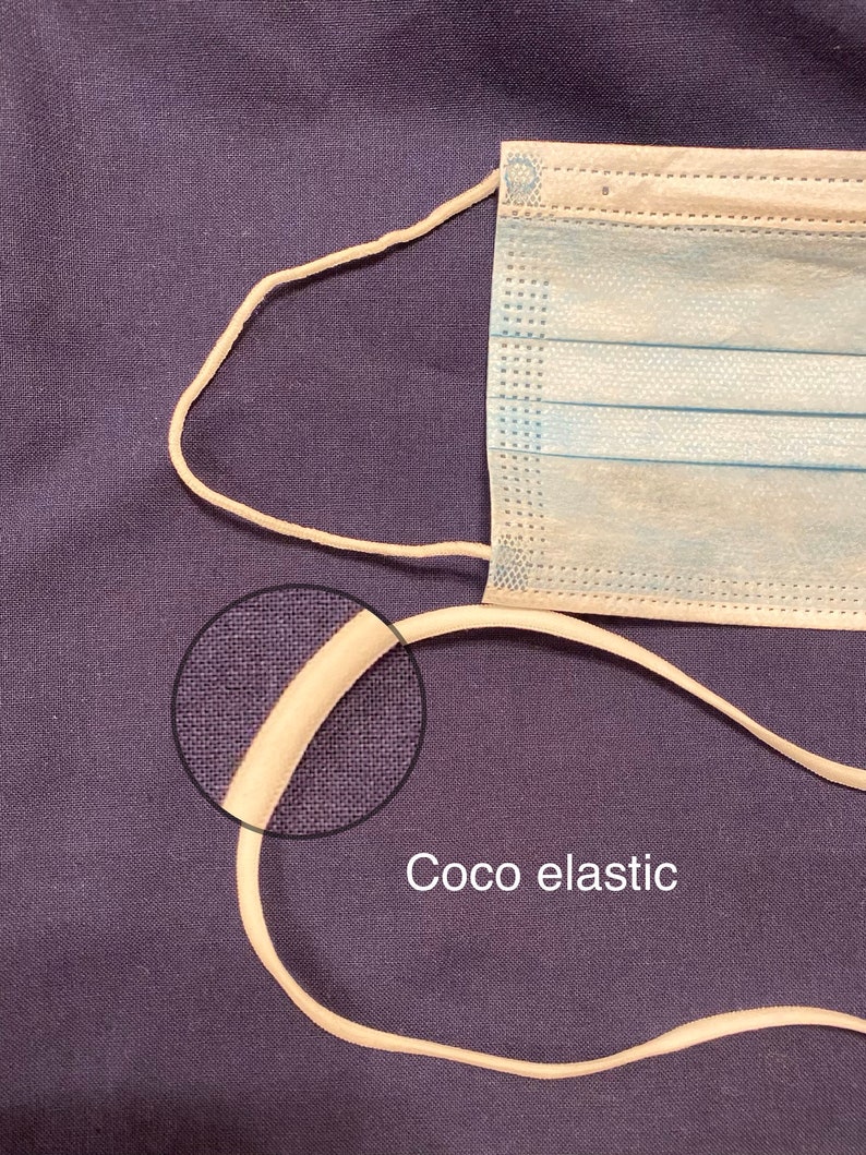 Pode incluir: Close-up de uma banda el&aacute;stica bege, tamb&eacute;m conhecida como "el&aacute;stico coco", sobre um fundo de tecido roxo. A banda el&aacute;stica &eacute; grossa e tem uma textura lisa.