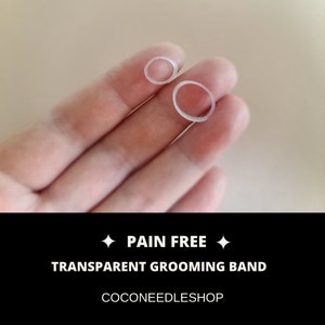 May include: Two clear plastic hair ties, one larger and one smaller, are held in a hand. The text "PAIN FREE" and "TRANSPARENT GROOMING BAND" is displayed below the image. The text "COCONEEDLESHOP" is displayed below the text.