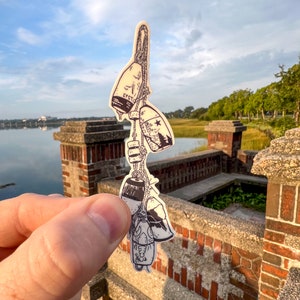 May include: A sticker featuring a cluster of nautical buoys, each with unique designs and markings. The buoys are cream-colored with black line art, connected by a rope. The background shows a scenic view of water and greenery.