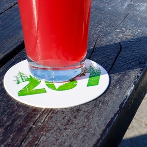 May include: A red beverage in a clear glass sits on a white round coaster with green text that reads "201" and a green tree design.