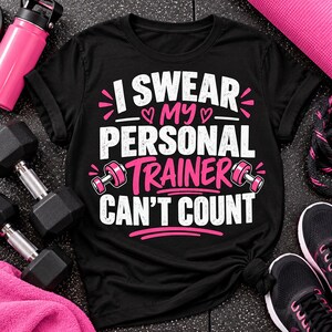 May include: A black short-sleeved t-shirt featuring white and pink text that reads I SWEAR MY PERSONAL TRAINER CAN'T COUNT. The design includes two small pink dumbbells and pink heart accents, centered on the front of the garment.