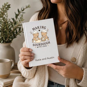 May include: A white hardback book titled Making Sourdough With Love, featuring a graphic of two brown teddy bears wearing chef hats and aprons kneading dough. The text Katie and Austin is printed in black at the bottom of the front cover.