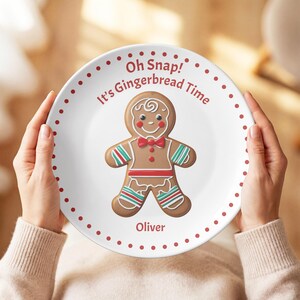Może przedstawiać: Biały ceramiczny talerz z centralną ilustracją ludzika z piernika z czerwoną muszką i paskami. Tekst Oh Snap! It's Gingerbread Time znajduje się nad postacią, a imię Oliver poniżej, całość otoczona jest obwódką z czerwonych kropek.