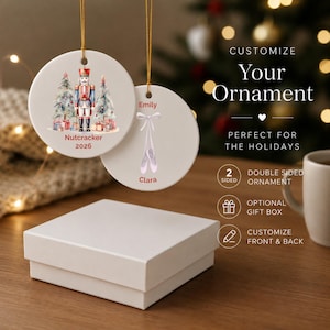 Może przedstawiać: Dwie okrągłe, kremowe ozdoby choinkowe ze złotymi sznurkami do zawieszania. Jedna przedstawia Dziadka do Orzechów, choinki i napis "Nutcracker 2026". Druga ma baletki i imiona "Emily" i "Clara."