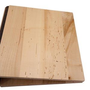 Pode incluir: Uma pilha de tábuas de madeira inacabadas. As tábuas são de cor clara com grão de madeira visível e pequenas manchas escuras. As bordas das tábuas são marrons escuras.