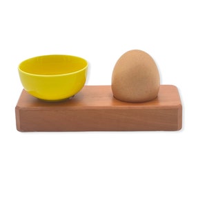Może przedstawiać: Drewniany stojak na jajka z żółtą ceramiczną miską i brązowym jajkiem. Stojak wykonany jest z naturalnego drewna i ma prosty design.