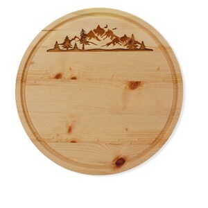 Könnte beinhalten: Rundes Holzbrett zum Schneiden mit einem eingravierten Berg- und Baummotiv am oberen Rand. Das Brett ist aus hellem Holz und hat eine natürliche Oberfläche.