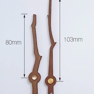 Op de afbeelding: Twee houten wijzers met een bruine afwerking en gouden accenten. De langere wijzer meet 103 mm en de kortere wijzer meet 80 mm.