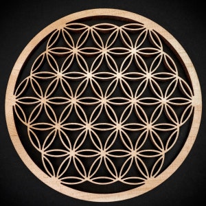 Può includere: Opera d'arte murale circolare in legno con il motivo del Fiore della Vita. Il legno chiaro è tagliato al laser per rivelare un motivo di cerchi sovrapposti, creando un motivo floreale simmetrico. L'opera ha un diametro di circa 30 cm ed è su sfondo scuro.