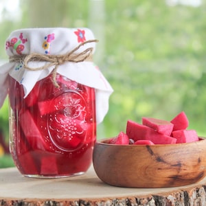 Peut inclure: Un bocal en verre rempli de betteraves roses marinées, recouvert d'un tissu blanc et d'une ficelle. Un bol en bois rempli de tranches de betteraves roses repose sur une souche d'arbre.