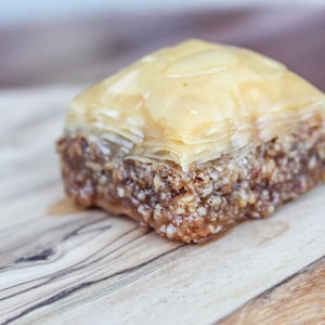 May include: A square piece of baklava, a traditional Greek pastry, with layers of flaky phyllo dough, a sweet filling, and a drizzle of honey or syrup. The baklava is sitting on a wooden cutting board.