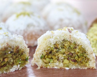 Op de afbeelding: Close-up van een pistachekoekje, doormidden gesneden, met een groene pistachevulling. Het koekje is bestoven met poedersuiker en omringd door andere koekjes en pistachekruimels.