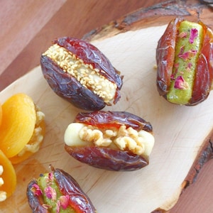 May include: A wooden cutting board with five dates filled with different toppings. The dates are filled with pistachio paste, sesame seeds, walnuts, and cream cheese. There are also dried apricots on the board.