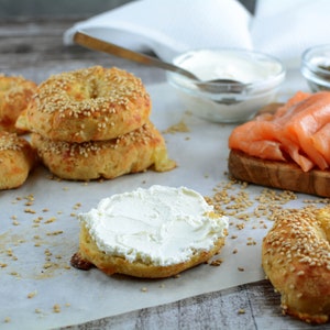 Op de afbeelding: Een close-up van goudbruine bagels, sommige met sesamzaad, met roomkaas, gerookte zalm, kappertjes en een kom roomkaas. De opstelling suggereert een ontbijt- of brunchthema.