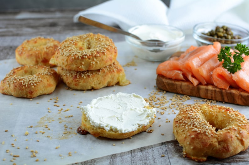 Op de afbeelding: Een close-up van goudbruine bagels, sommige met sesamzaad, met roomkaas, gerookte zalm, kappertjes en een kom roomkaas. De opstelling suggereert een ontbijt- of brunchthema.