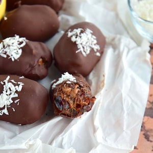 Op de afbeelding: Close-up van chocolade bedekte dadels die bestrooid zijn met geraspte kokosnoot. De dadels zijn gerangschikt op wit perkamentpapier. Eén dadel is gedeeltelijk opgegeten, waardoor de bruine dadelvulling zichtbaar wordt.