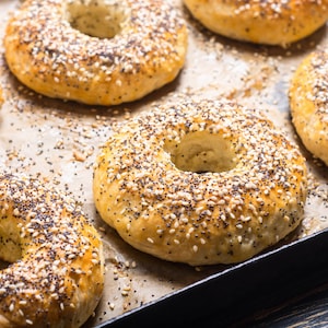 Op de afbeelding: Close-up van versgebakken bagels op een bakplaat. De bagels zijn goudbruin, met sesamzaad en maanzaad. De bagels zijn gerangschikt op bakpapier.