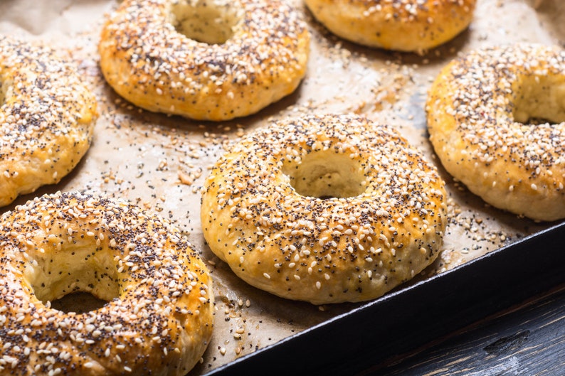 Op de afbeelding: Close-up van versgebakken bagels op een bakplaat. De bagels zijn goudbruin, met sesamzaad en maanzaad. De bagels zijn gerangschikt op bakpapier.