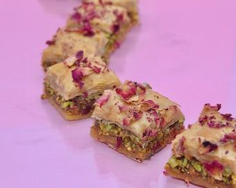 May include: A close-up of a row of baklava pastries topped with rose petals. The pastries are arranged in a curved line on a pink surface.