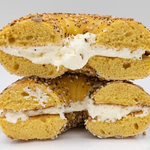 Op de afbeelding: Een close-up van een in plakken gesneden bagel gevuld met roomkaas. De bagel is goudbruin, bedekt met zaden en doorgesneden om de romige vulling te onthullen. De bagel is op een andere helft gestapeld.