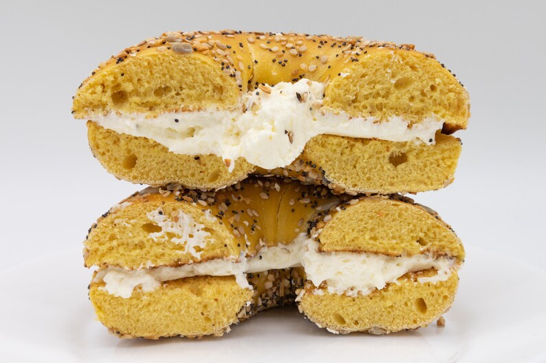Op de afbeelding: Een close-up van een in plakken gesneden bagel gevuld met roomkaas. De bagel is goudbruin, bedekt met zaden en doorgesneden om de romige vulling te onthullen. De bagel is op een andere helft gestapeld.