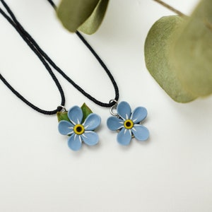 Puede incluir: Dos delicados collares con colgantes de flores de nomeolvides. Las flores son azules con centros amarillos y hojas verdes. Los collares son cordones negros.