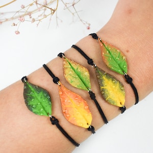 Puede incluir: Cinco pulseras con bandas elásticas negras y dijes en forma de hoja. Las hojas están hechas de arcilla polimérica y están pintadas en tonos de verde, amarillo y naranja. Cada hoja tiene una superficie texturizada que se asemeja a gotas de agua.