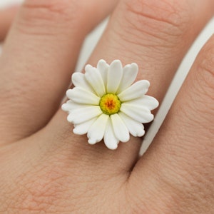 May include: A white daisy ring with a yellow centre. The ring is worn on a person's finger.
