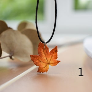 May include: A necklace with a pendant shaped like an orange and brown maple leaf. The necklace is made with a black cord.