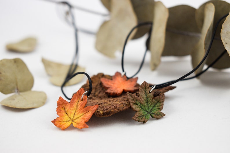 May include: Three necklaces with leaf pendants. The leaves are made of polymer clay and are painted in shades of orange, green, and brown. The necklaces are made with black cord.