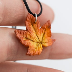 May include: A necklace with a fall-colored maple leaf pendant. The leaf is made of polymer clay and is painted in shades of orange, yellow, and brown.