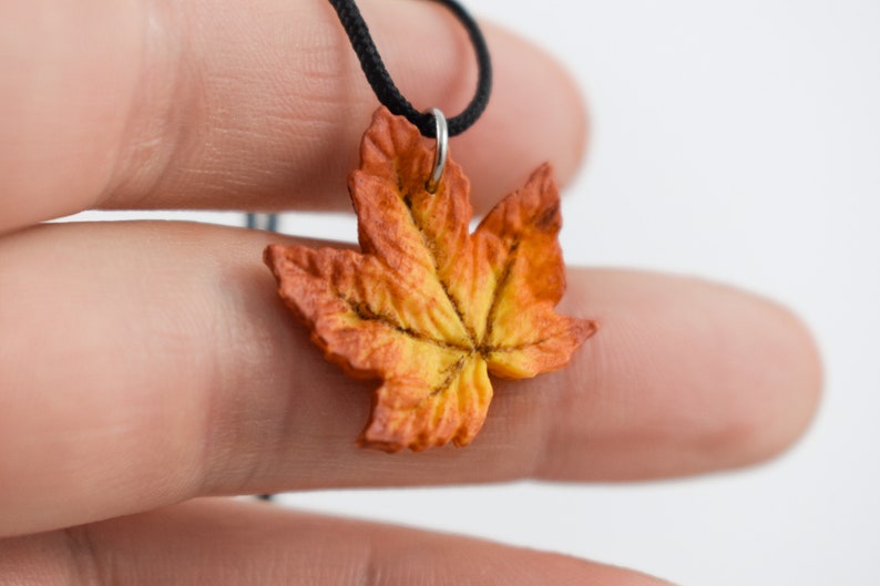 May include: A necklace with a fall-colored maple leaf pendant. The leaf is made of polymer clay and is painted in shades of orange, yellow, and brown.