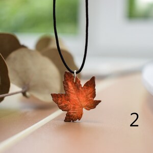May include: A necklace with a copper maple leaf pendant. The leaf is a deep orange color and has a detailed design. The necklace is on a black cord.