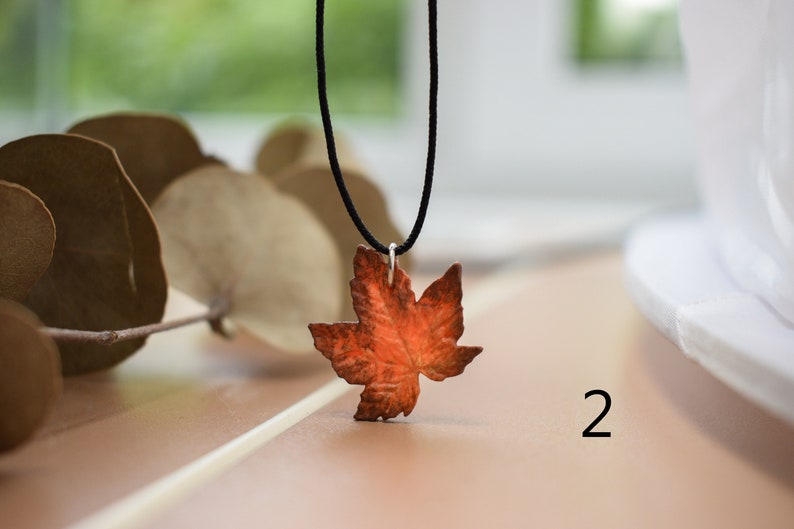 May include: A necklace with a copper maple leaf pendant. The leaf is a deep orange color and has a detailed design. The necklace is on a black cord.