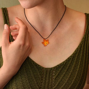 May include: A woman wearing a green knitted tank top with a black cord necklace featuring a fall-colored maple leaf pendant.