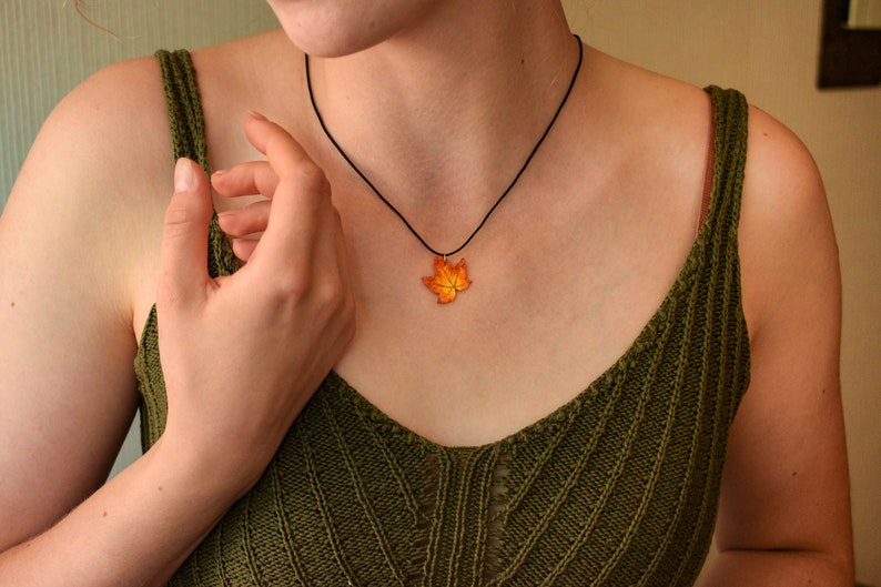 May include: A woman wearing a green knitted tank top with a black cord necklace featuring a fall-colored maple leaf pendant.