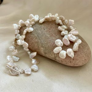 Puede incluir: Un brazalete de perlas con cierre plateado. El brazalete está compuesto por perlas blancas cremosas de forma irregular. Las perlas están dispuestas en un patrón fluido, creando un aspecto natural y elegante. El brazalete se exhibe sobre una piedra lisa de color marrón claro.