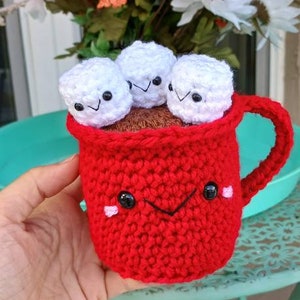 May include: A red crocheted mug with a smiling face and three white crocheted marshmallows inside. The mug has a handle and is sitting on a green table.